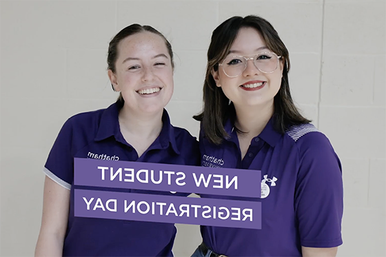 两名身穿紫色制服的学生志愿者一起微笑拍照. 他们面前的紫色横幅上写着“新生注册日”.